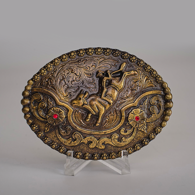 The Ruby Bull Rider Buckle is an ornate, oval bronze piece with an embossed horse amid intricate floral and swirl designs, echoing Texas-style buckles. Displayed on a clear stand against a simple backdrop, it exudes rustic charm and craftsmanship.