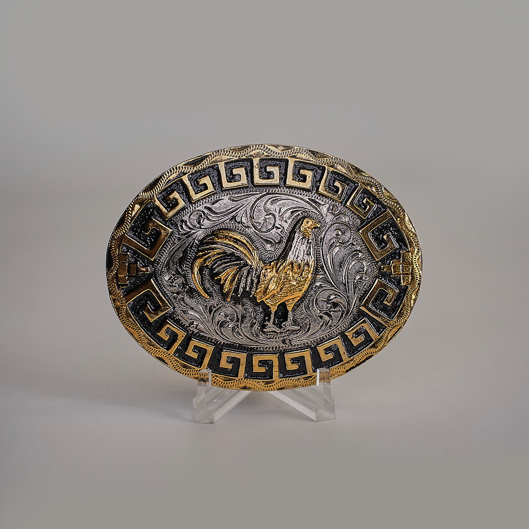 The Charro Rooster Buckle is an oval belt buckle with a western design, featuring a gold rooster on a silver background with swirling patterns. It has a Greek key motif in gold and black on the border. Made from alpaca silver, it is displayed on a clear stand against a gray background.