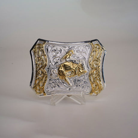 The Spade Bull Rider Buckle is a Texas-style piece crafted from alpaca silver. It features intricate engravings of a cowboy riding a bull, with silver and gold accents that pop against its plain background, showcasing the detailed western design.