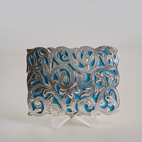 The Charro Turquoise Buckle is an ornate alpaca silver piece featuring intricate scrolling patterns on a vibrant blue background. Its cut-out design showcases exceptional craftsmanship with highlighted curves and flourishes, reminiscent of classic Texas-style buckles, displayed upright on a stand.