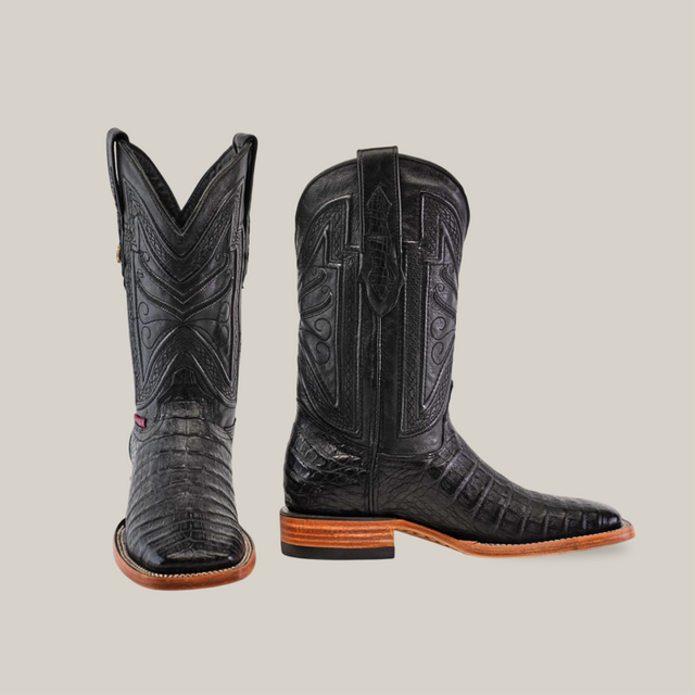 Exotic Caiman Belly boots in black feature intricate stitching for cowboy enthusiasts. Their square toe design and glossy caiman belly finish are complemented by wooden soles with a subtle heel. One boot is upright, the other is side-on to showcase their unique style.