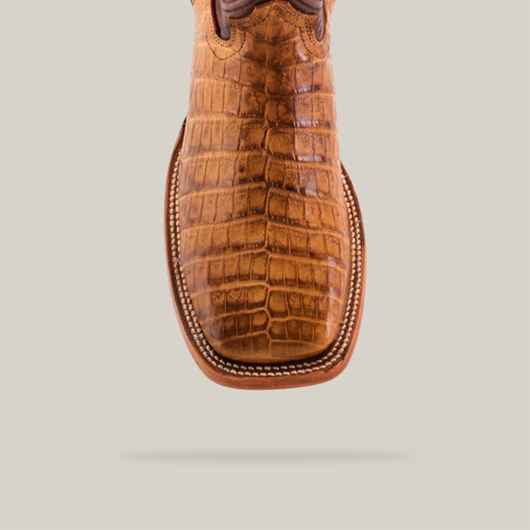 Close-up of the Exotic Caiman Belly Boot in Camel features brown, crocodile-patterned leather with a square toe design and detailed white stitching. Set against a light beige background.