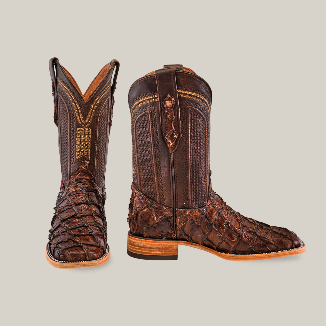 The Exotic Piraruco Fish Brown boots feature a scaled texture made from dark brown pirarucu leather. Embracing Western fashion, they showcase intricate stitching on the upper shaft and a wooden heel. One boot is upright; the other angles slightly to display its side detail.