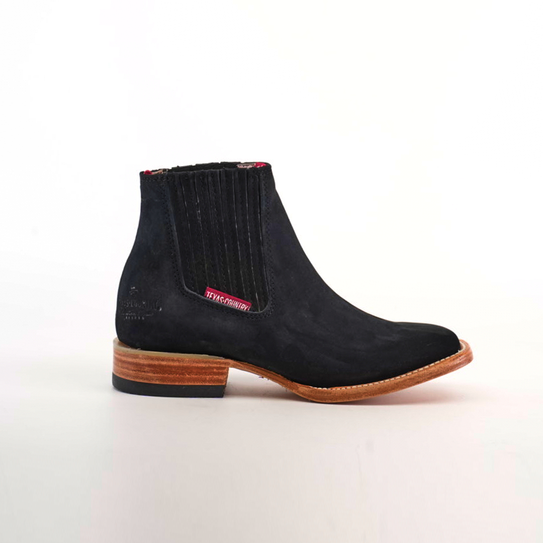 The Prime Suede Black Square Toe boots feature black suede with a wooden heel, contrasting red tag by the elastic panel, smooth finish, stitched details, and a slightly elevated sole. The design subtly nods to cowboy fashion against a white background.