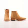 The Prime Suede Honey Square Toe boots are tan, ankle-length, and feature elastic side panels, a square toe, and light-colored soles. Displayed on a white background, they evoke cowboy style with one boot facing forward and the other to the side.