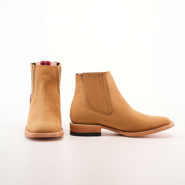 The Prime Suede Honey Square Toe boots are tan, ankle-length, and feature elastic side panels, a square toe, and light-colored soles. Displayed on a white background, they evoke cowboy style with one boot facing forward and the other to the side.