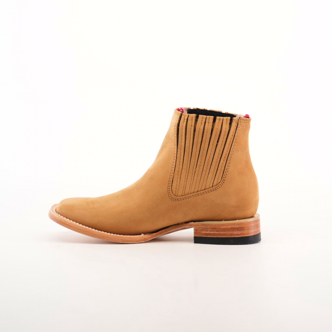 The Prime Suede Honey Square Toe is a tan leather Chelsea boot featuring a wooden heel and black rubber sole, with a hint of cowboy style. It has elastic side panels for easy slip-on and a square toe for a modern look, set against a plain white background.