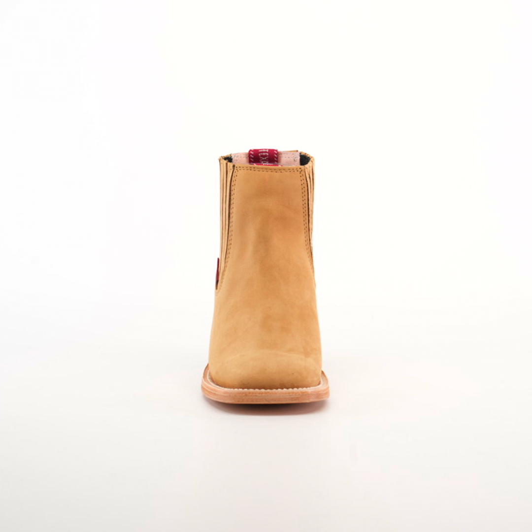 The Prime Suede Honey Square Toe is showcased against a white background. Featuring a tan suede hue, this ankle boot has a flat sole and square toe. A red plaid and beige pull tab adds contrast, embodying the Cowboy Fashion trend.