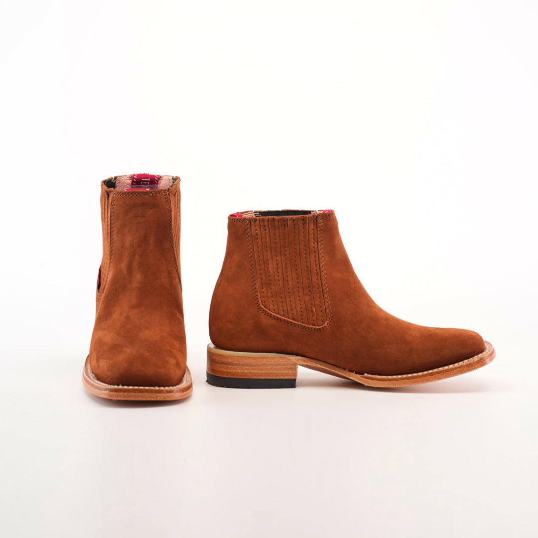 The Prime Suede Brick Square Toe boots feature brown suede with flat heels, red plaid lining peeking from the top, and elastic side panels for easy slip-on. One boot faces forward while the other displays its side profile in a classic cowboy style.