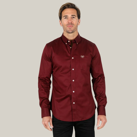 A man confidently stands before a light gray backdrop, donning the stylish and comfortable Men’s Single Pocket Logo Modern Fit Stretch Dress Shirt in Burgundy (CTL9260), featuring a button-down collar and logo on the chest pocket. He gazes directly at the camera, exuding confidence.