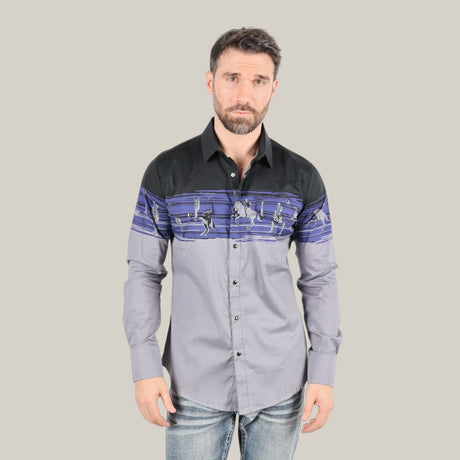 A bearded man wears the Modern Fit Cotton Stretch Rodeo Panoramic Print Shirt - AZL9941, featuring a dark upper section with a colorful geometric pattern and a lighter lower section. Standing against a plain backdrop paired with jeans, he stylishly gazes at the camera.