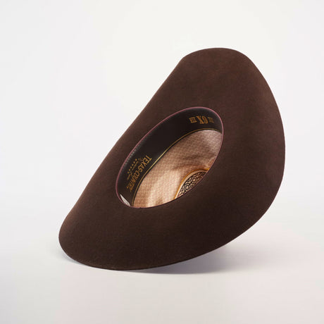 The 6X Country Brown hat, a wide-brimmed felt creation from premium sheep wool, is artfully displayed. Its interior features a patterned lining with TEXANA and XX, embodying Western classic style. The display tilts to reveal both exterior and interior details against a plain background.