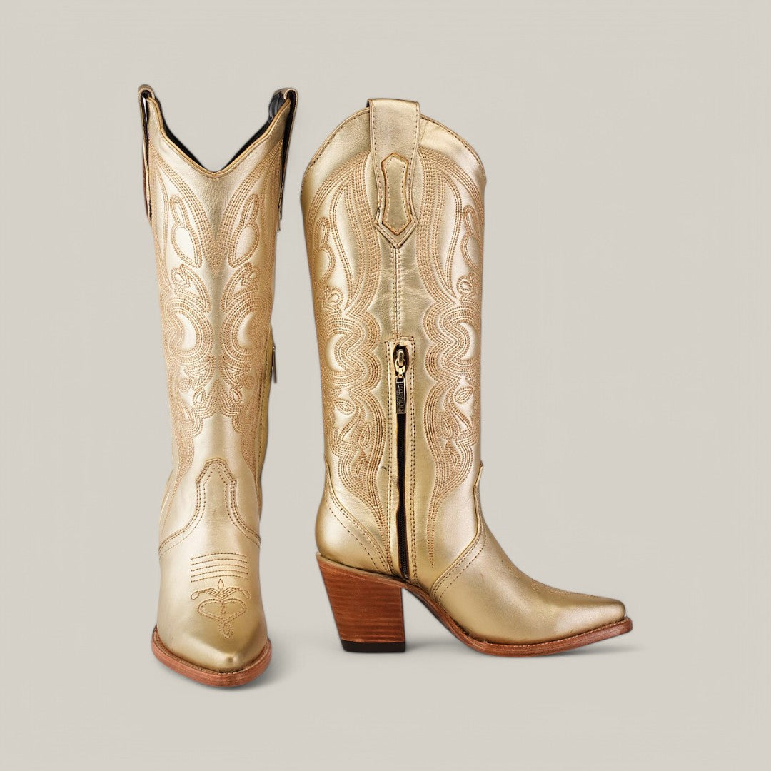 Introducing the Linda Tall Shaft Gold cowboy boots from our Platinum Collection, featuring intricate stitching, a sleek side zipper, and stacked wooden heels. One boot stands upright while the other tilts forward to showcase its pointed J Toe design.