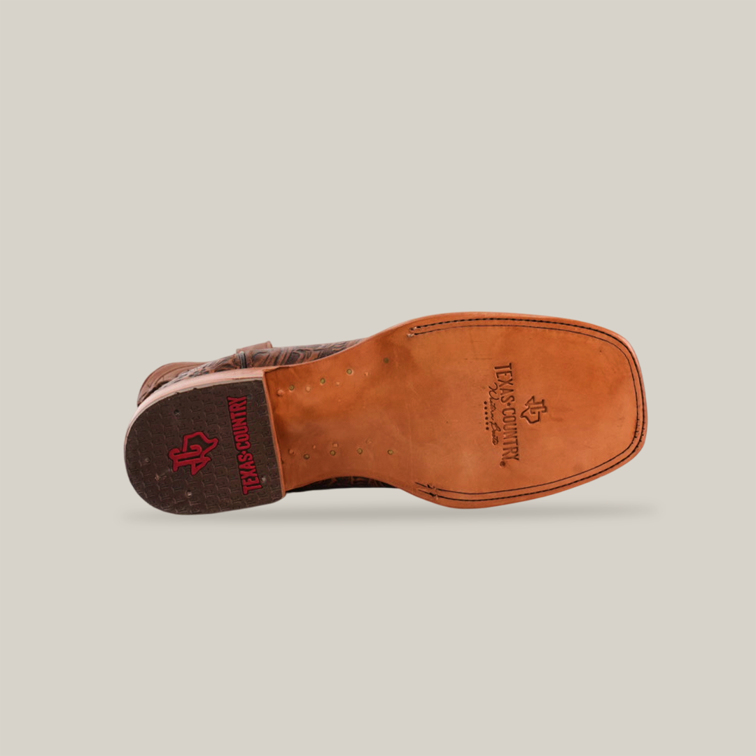 The image features the sole of the Handtooled Leave Brown - Square Toe cowboy boot. Made from premium cowhide leather, its tan sole displays an engraved Texas Country logo in red and is accented with small metal studs, highlighting the craftsmanship.