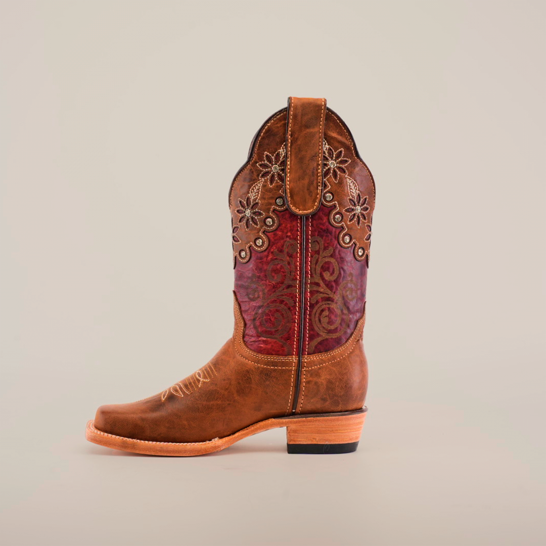 The Grecas Orix boot features a brown and reddish leather exterior with intricate floral embroidery on the shaft, a scalloped collar, and a narrow square toe. It has a wooden heel and is showcased against a plain white background.