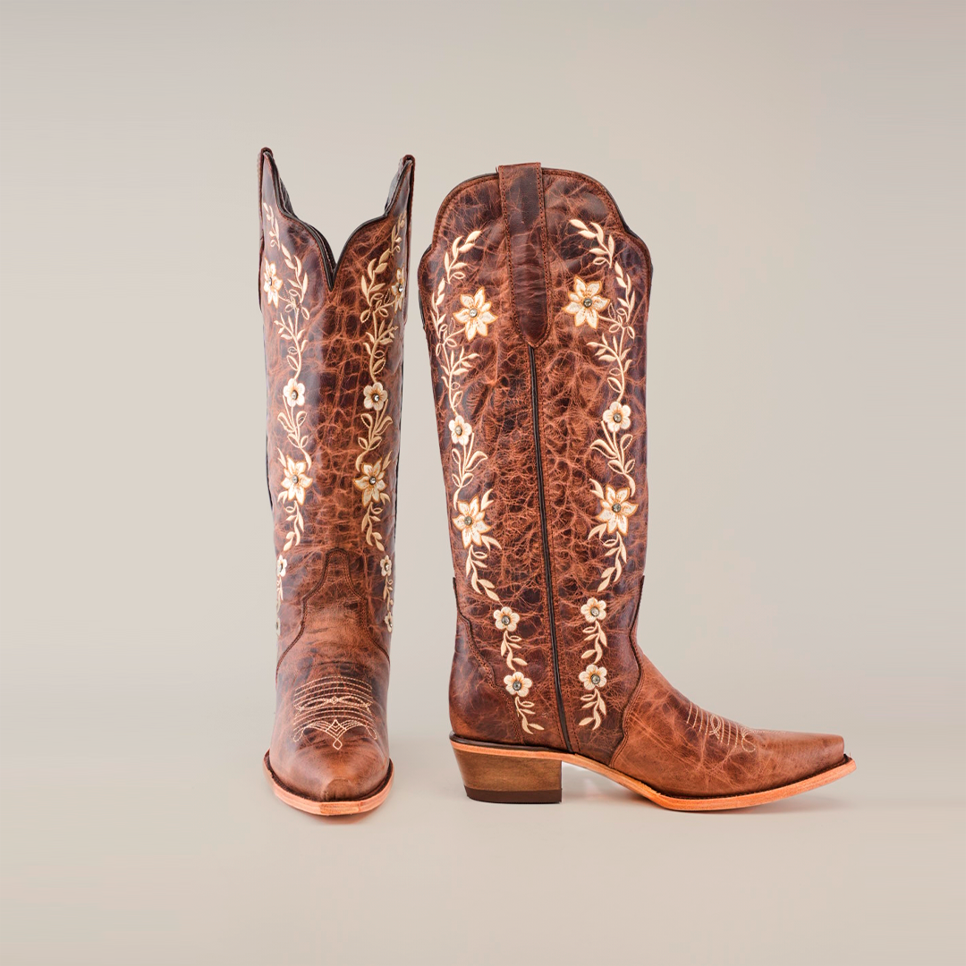 The Mia Brown Tall Shaft Snip Toe boots, crafted from premium leather, feature intricate white floral embroidery. One boot stands upright and the other is angled forward, highlighting the design. These handcrafted boots have pointed toes and low heels against a plain background.