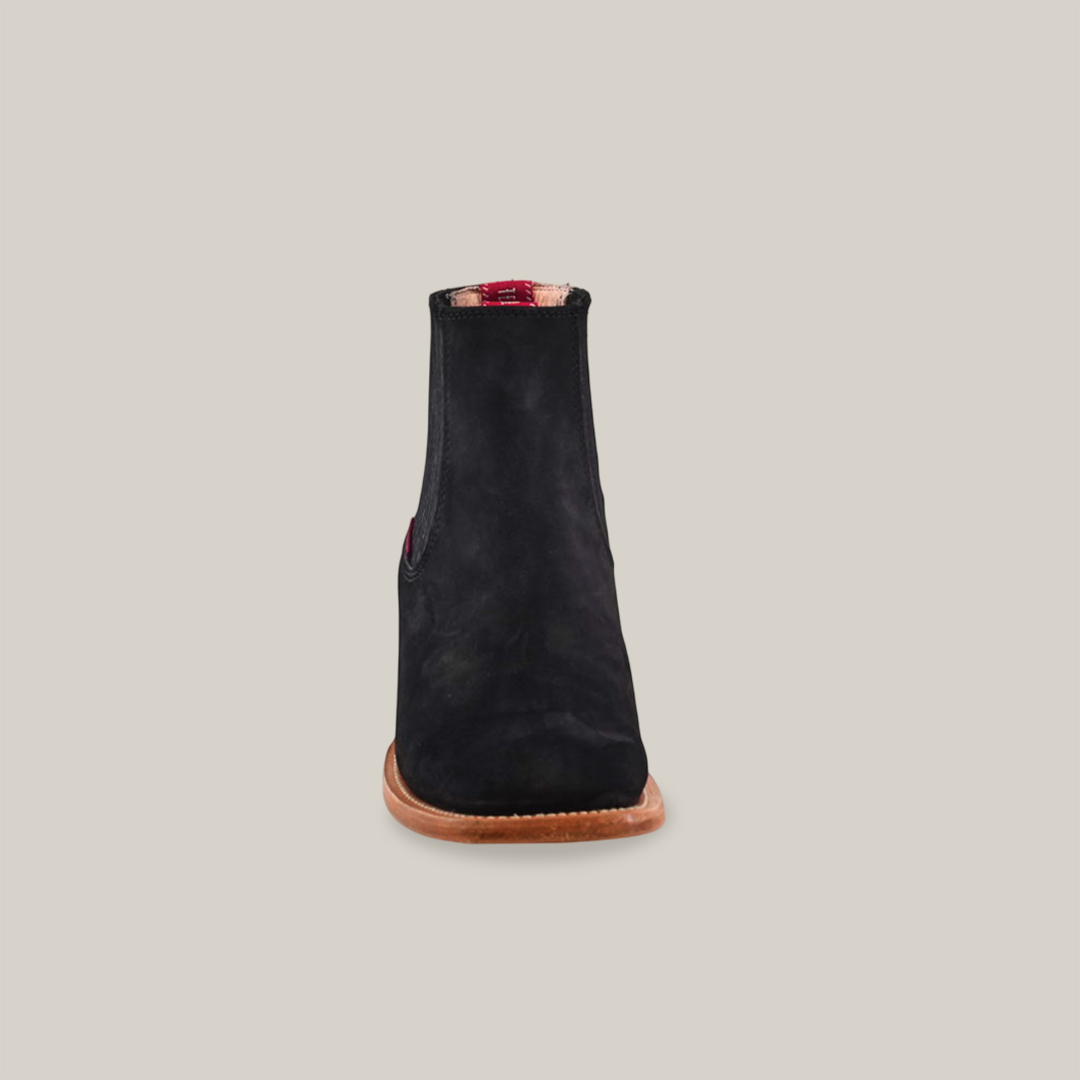 The Prime Suede Black - Square Toe is a chic black suede ankle boot with a flat brown sole, standing upright against a white background. Its interior showcases a red plaid pattern, highlighting premium materials for comfort and style.