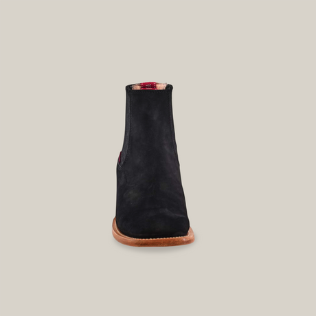 The Prime Suede Black - Square Toe is a chic black suede ankle boot with a flat brown sole, standing upright against a white background. Its interior showcases a red plaid pattern, highlighting premium materials for comfort and style.