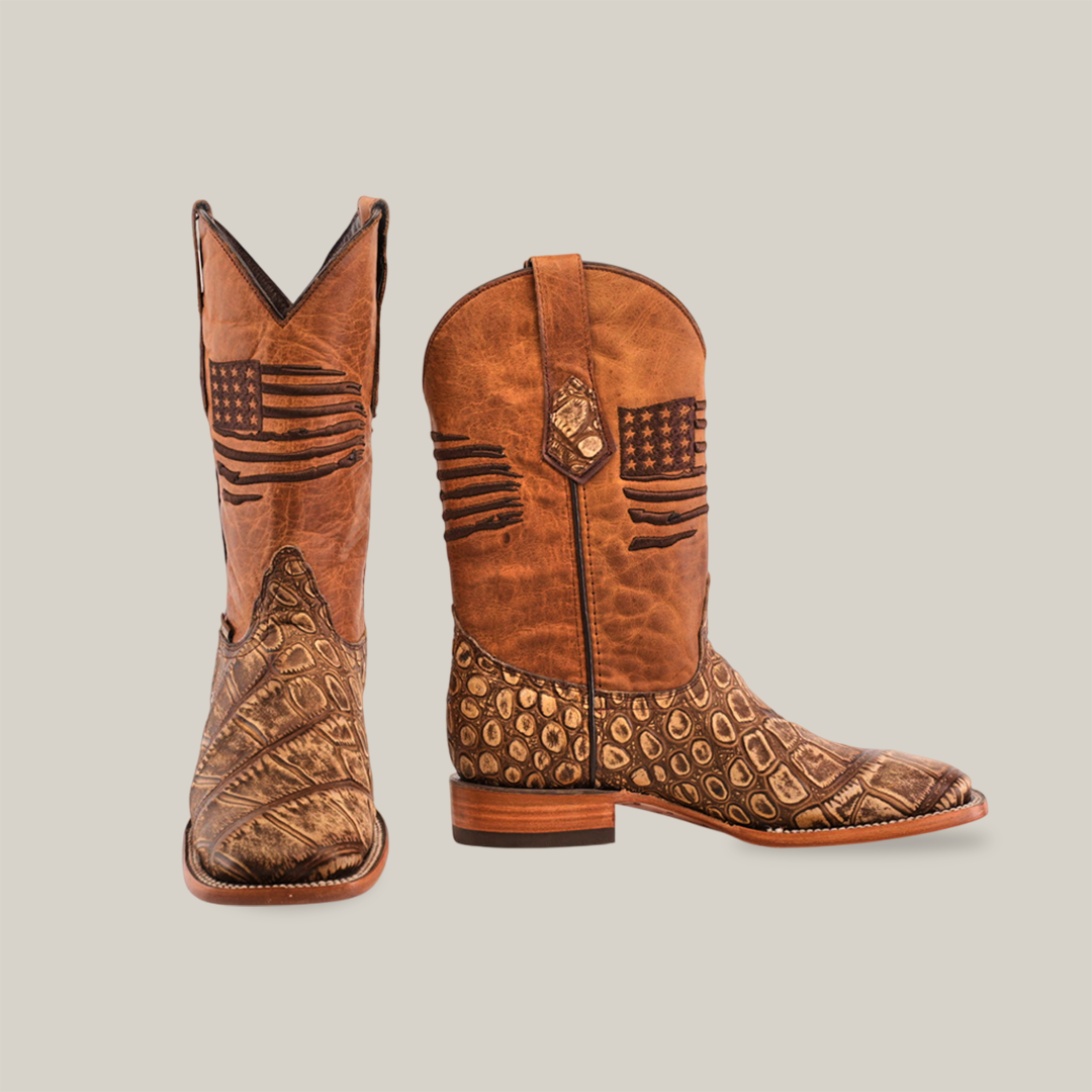 The American Alligator Print Rustic Orix boots feature a square toe with American flag designs on the shaft, leather soles, and brown textures with decorative patterns. One boot stands upright while the other tilts slightly forward.