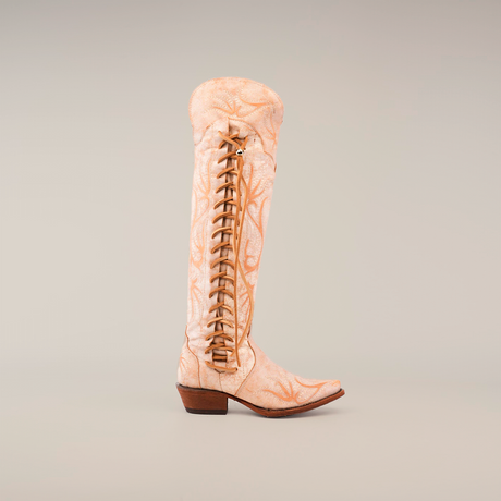 The Emma Alaska Orix-Tall Shaft - Snip Toe is a masterpiece knee-high boot, featuring tan suede, intricate crisscross leather laces, and a wooden heel. It is handcrafted from premium leather with a decorative star pattern and rounded toe against a white background.