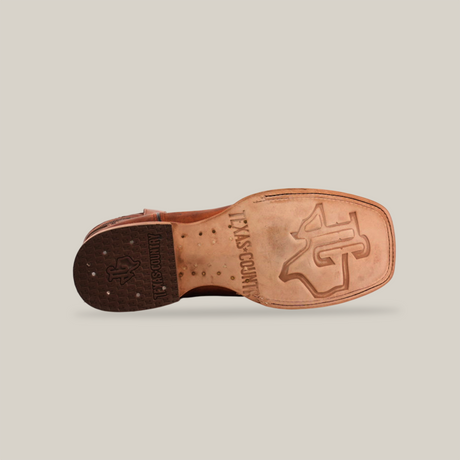 The Rustic Brown - Square Toe cowboy boots bottom view showcases a light brown sole with TEXAS COUNTRY and its logo. The heel, made of rustic brown cowhide leather, exhibits texture and branding against a plain white background while lying on its side.