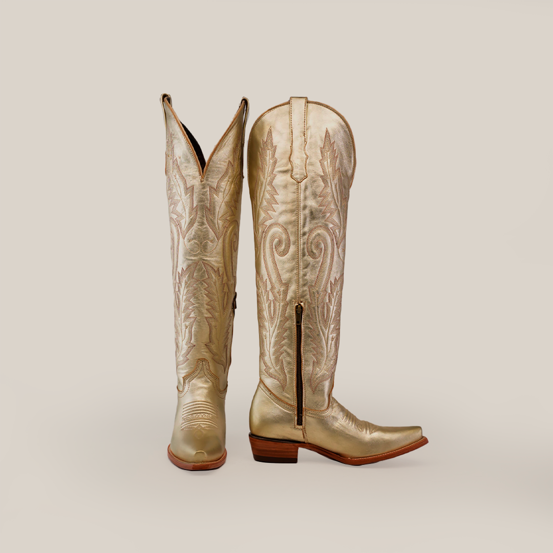 The Brittany Gold Tall Shaft Snip Toe boots feature intricate floral patterns on ornate leather, with one womens boot upright and the other showing a side zipper and pointed toe. Crafted with care, these gold treasures sit against a plain gray background with low wooden heels.