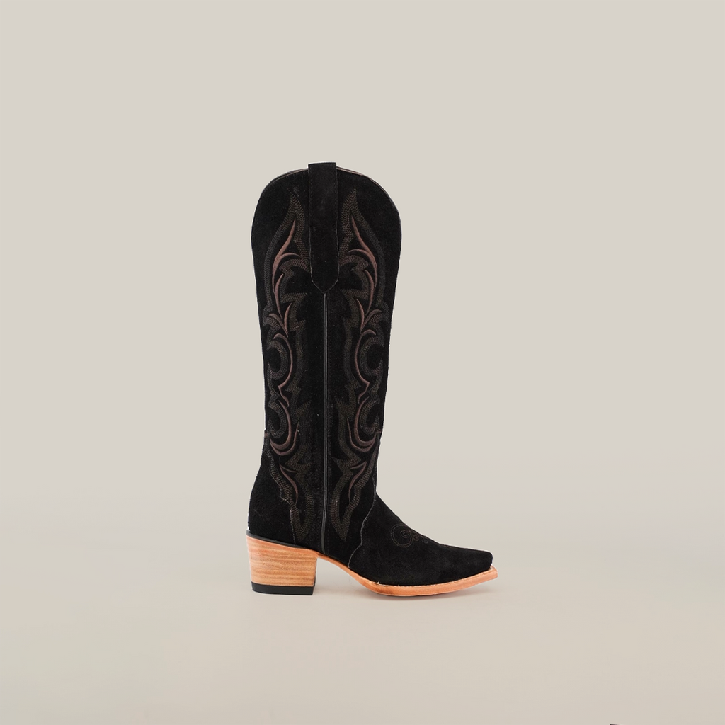 The Amara Black Suede - Tall Shaft - Snip Toe cowboy boot features intricate brown embroidery, a snip toe, and a wooden stacked heel. Its design is striking against the plain white background.