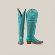 A pair of Brittany Rustic Ceramic Tall Shaft Snip Toe Boots in turquoise features intricate swirling embroidery, brown wooden heels, and a genuine leather sole. One boot stands upright while the other tilts, showing off the side zipper and pointed toe design.