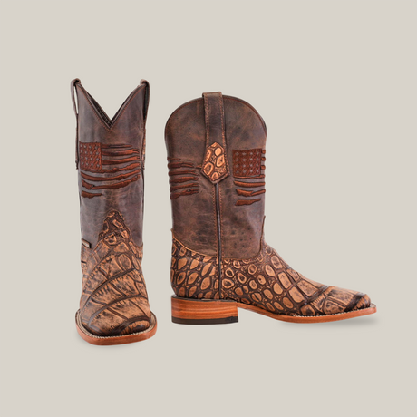 The American Alligator Print Rustic Brown boots feature a square toe design, combining smooth and textured brown leather with an embossed pattern. They display a detailed American flag near the top and have sturdy wooden heels, showcasing rustic handcrafted artistry.