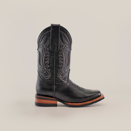 The Barcelona Black Slip Resistant Sole Rodeo Toe boot showcases intricate shaft embroidery, a brown wooden heel, and a stylish rodeo toe. Made from premium leather, it elegantly stands against a plain white background.