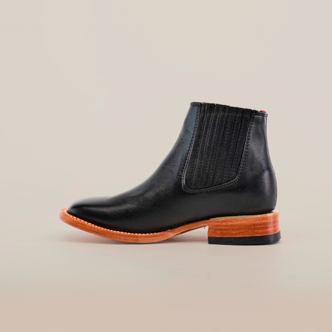 The Napa Black Square Toe ankle boot features sleek black leather, elastic side panels, and rounded toes. Its wooden sole and slight heel make it an essential addition to any fashion-forward wardrobe, showcased perfectly against a plain white background.