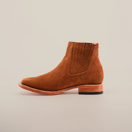 The Prime Suede Caramel Square Toe boots offer rustic charm with their brown suede finish, side elastic panel, decorative stitching, and square toe design. They feature a wooden sole paired with a black heel and are showcased on a plain white background.