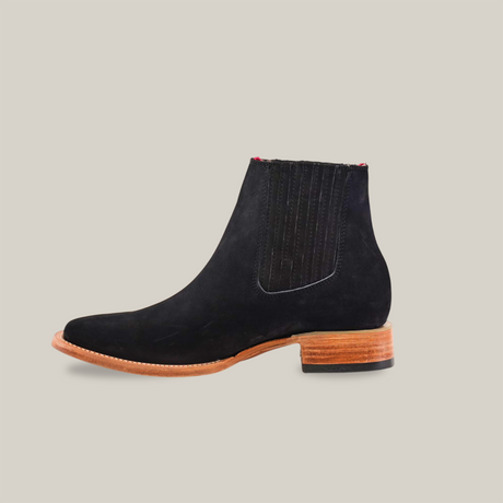 Side view of Prime Suede Black - Square Toe, a Chelsea boot in premium materials with a wooden sole and heel. It features elastic side panels and a slight point at the toe, echoing classic western style against a plain white background.