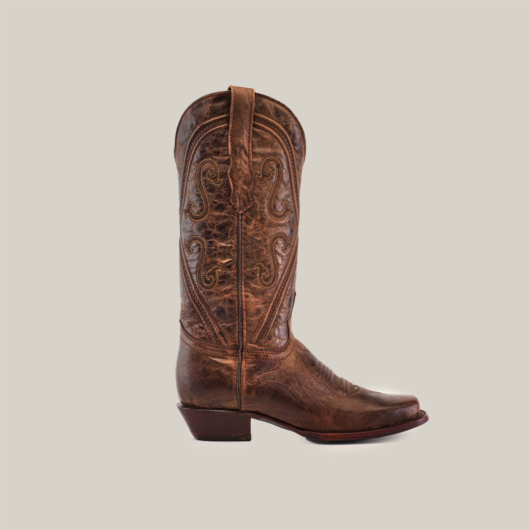 The Piola Tabaco boot is a brown leather cowboy boot with a narrow square toe, mid shaft, intricate stitching, and low heel, set against a plain white background.