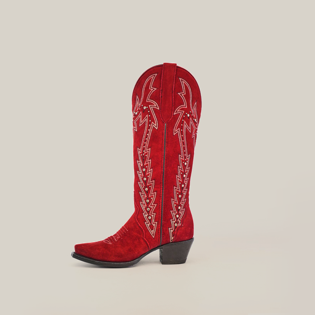 The Amara Red Suede Snip Toe boot, made from luxurious red suede leather with intricate white stitching, boasts an elegant design with a tall shaft and low heel, showcased against a plain white background.