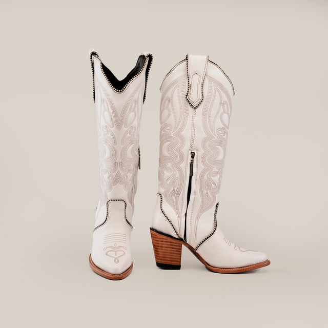 The Linda Studs Tall Shaft Frost White - J Toe cowboy boots, part of the Platinum Collection, boast artisanal craftsmanship with intricate embroidery. Made from premium leather, these stylish boots feature pointed toes, a side zipper, and stacked wooden heels against a plain background.