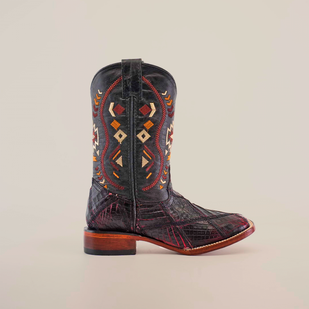 A single black Exotic American Alligator Patchwork boot displays intricate multicolored geometric stitching with red, brown, and white accents. It features a unique design and a low wooden heel, crafted in smooth leather with a hint of black cherry hue, standing upright on a white background.