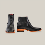 A pair of Napa Black Square Toe ankle boots showcases its leather build, elastic side panels, and wooden stacked heels. One boot faces forward while the other shows a side view, emphasizing its handcrafted design and texture reminiscent of a refined cowboy scene.