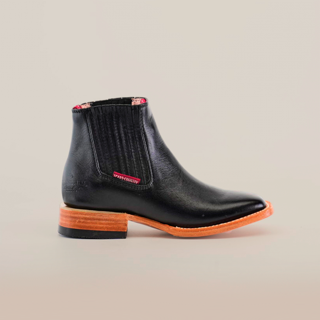 The Napa Black Square Toe boot features black Napa leather with a square toe design, brown wooden sole, slight heel, side elastic panels, and a decorative red tag on plain white background. Perfect addition to the fashion-forward wardrobe.