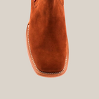 Close-up of the front of the handcrafted Prime Suede Brick Square Toe Ankle Boot. The brown leather shoe, with a cowboy-inspired design, highlights exceptional craftsmanship featuring visible stitching around the sole and an upper material that resembles suede.