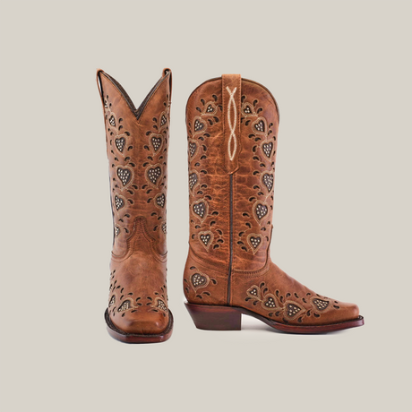 The Studded Hearts Orix boots feature premium leather with intricate heart patterns and cut-outs. One boot stands tall while the other leans slightly, highlighting artisan craftsmanship with a stitched XO design at the top.