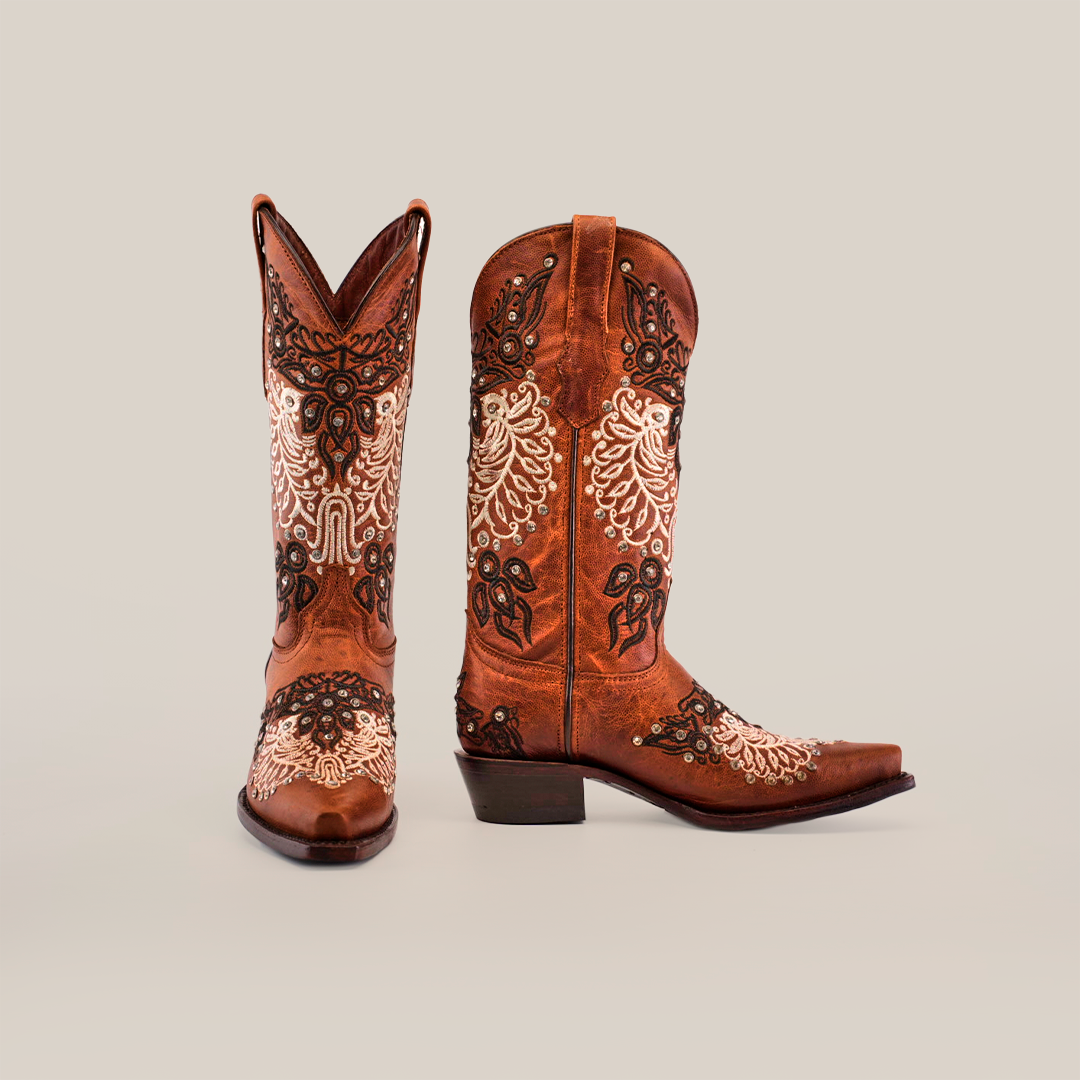 The Faenza Crystals Tabaco boots, with a mid shaft and snip toe, showcase intricate white floral and black swirl embroidery on a plain white background. One boot is angled slightly forward, highlighting their stunning design.