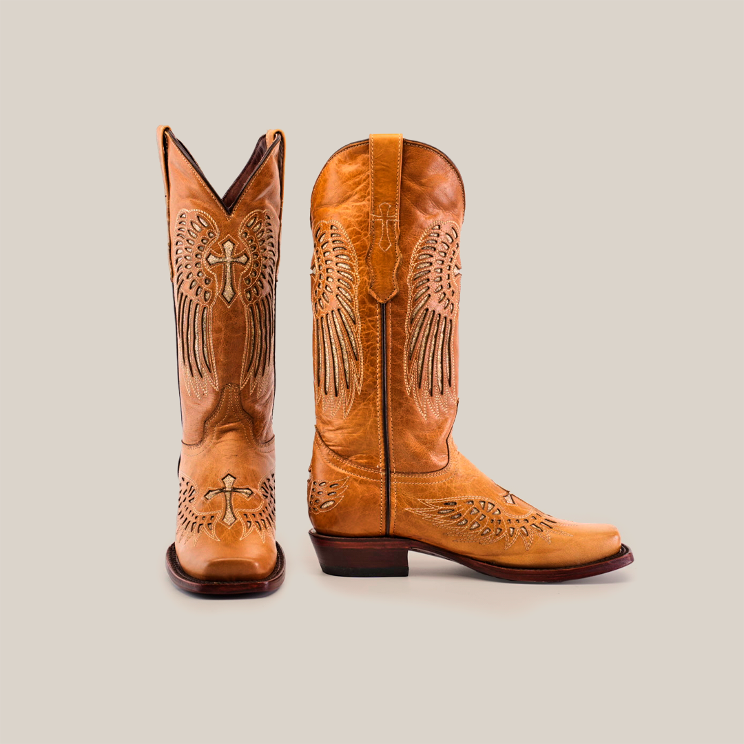 A pair of premium leather boots, the Texas Cross Glitterbomb Orix - Mid Shaft - Narrow Square Toe, with tan cowboy style and intricate wing and cross embroidery. One boot stands upright while the others sole and back are highlighted at an angle.