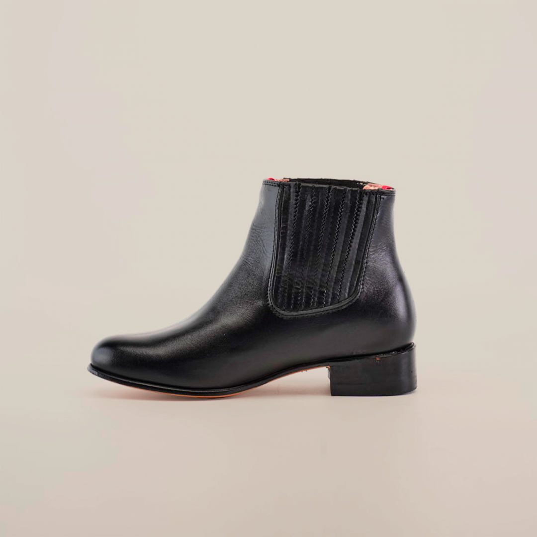 Introducing the Napa Black Square Toe Ankle Boot: a chic black leather design with an elastic side panel for easy slip-on and a subtle red outline on the sole, set against a plain white background, perfect for the fashion-forward wardrobe.