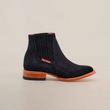 The Prime Suede Black Square Toe boots feature black suede with a wooden heel, contrasting red tag by the elastic panel, smooth finish, stitched details, and a slightly elevated sole. The design subtly nods to cowboy fashion against a white background