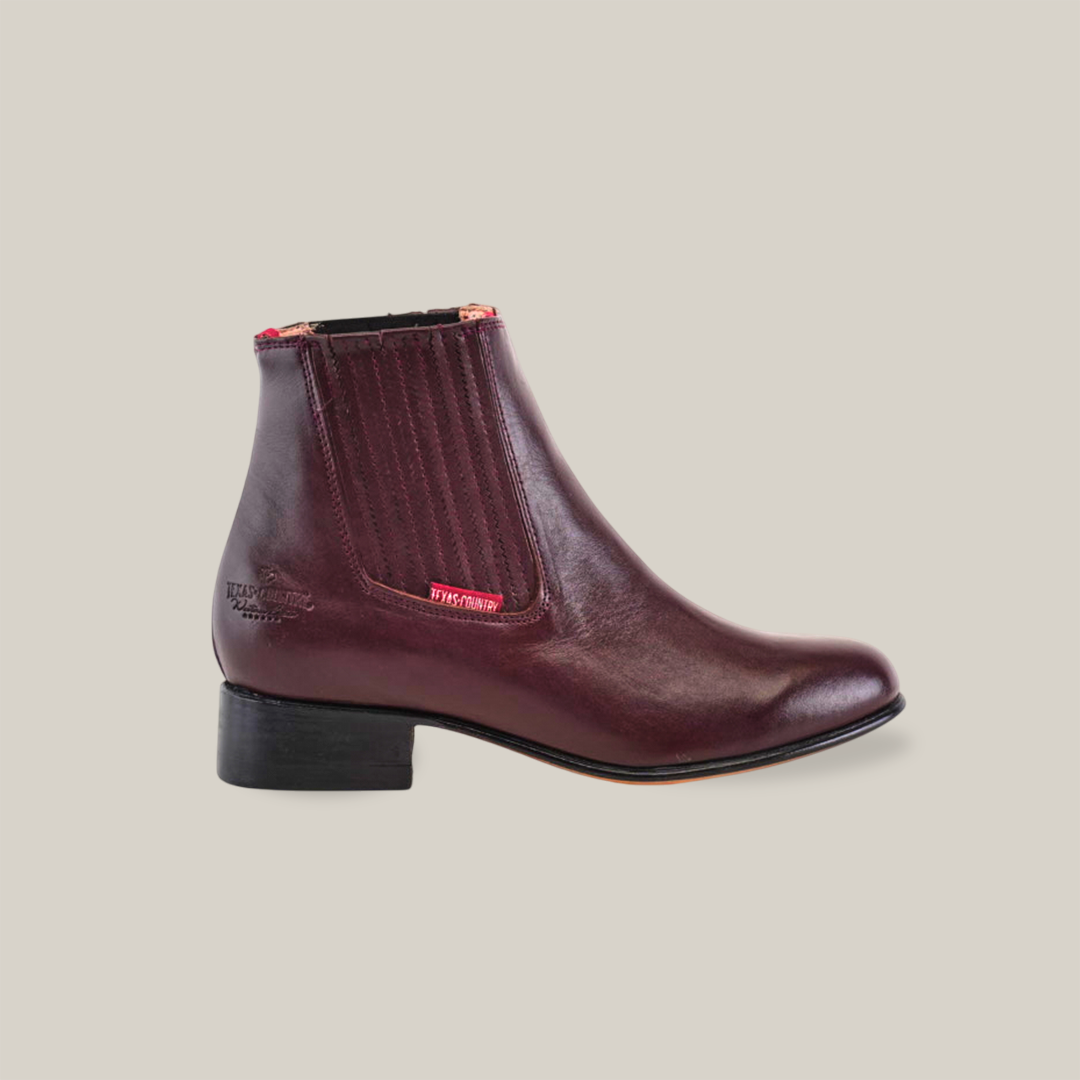 The Napa Wine - Round Toe is a leather ankle boot in deep burgundy with elastic side panels, a low black heel, and red stitching near the top. HEY DUDE adorns the side, showcasing its artesanía excepcional for those who appreciate style.