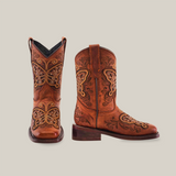 The Butterfly Studs Cogñac boots are crafted from cowhide leather with intricate butterfly embroidery and swirling patterns. They feature a square toe, classic Western style, pull tabs, and a low heel, all set against a plain white background.