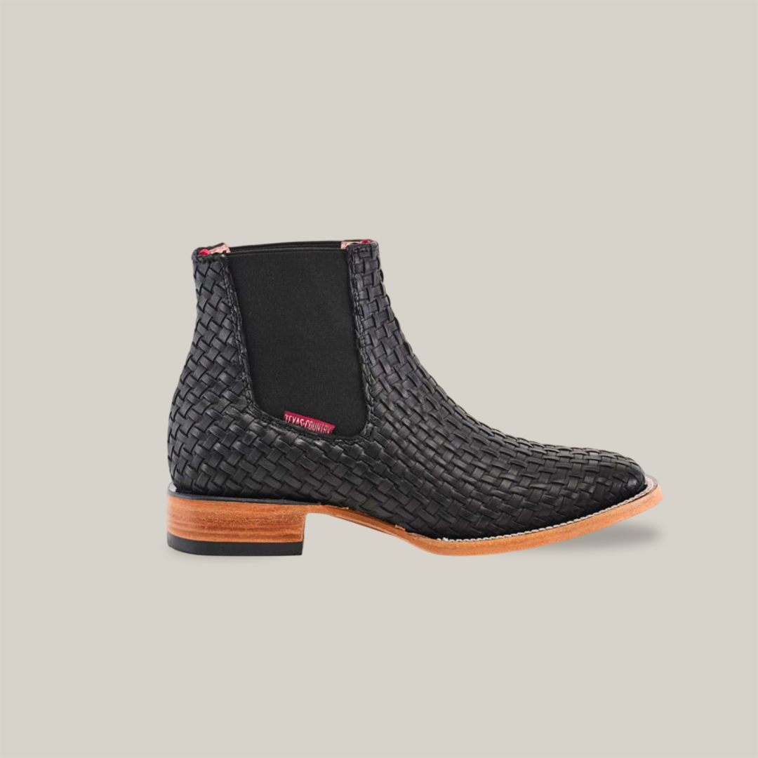 The Basket Weave Black Square Toe ankle boot features a chic basket weave design, square toe, brown wooden heel and sole, black elastic side panels, and a playful pink back tab. Set on a crisp white background, it exudes style and sophistication.