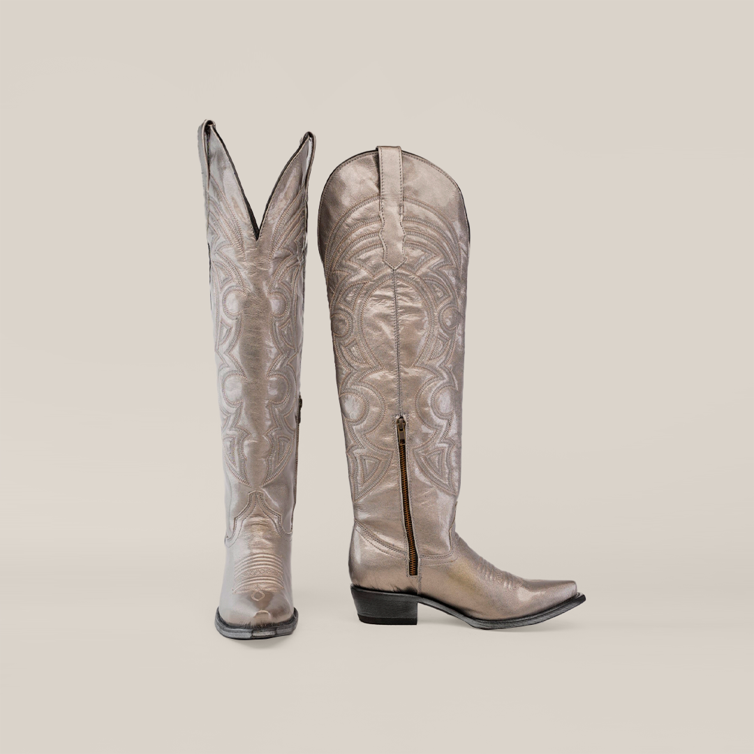 Two Taylor Mercury - Tall Shaft - Snip Toe boots, made of shiny metallic material with intricate swirling patterns. One stands upright, and the other is angled to show a side zipper and leather sole, set against a light gradient background.