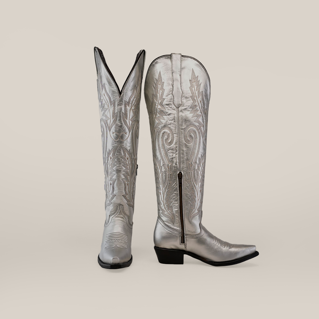 A pair of Brittany Silver Tall Shaft Snip Toe Boots, expertly crafted from premium leather with detailed stitching and side zippers. One boot stands upright while the other leans on a plain backdrop, highlighting their handcrafted elegance.